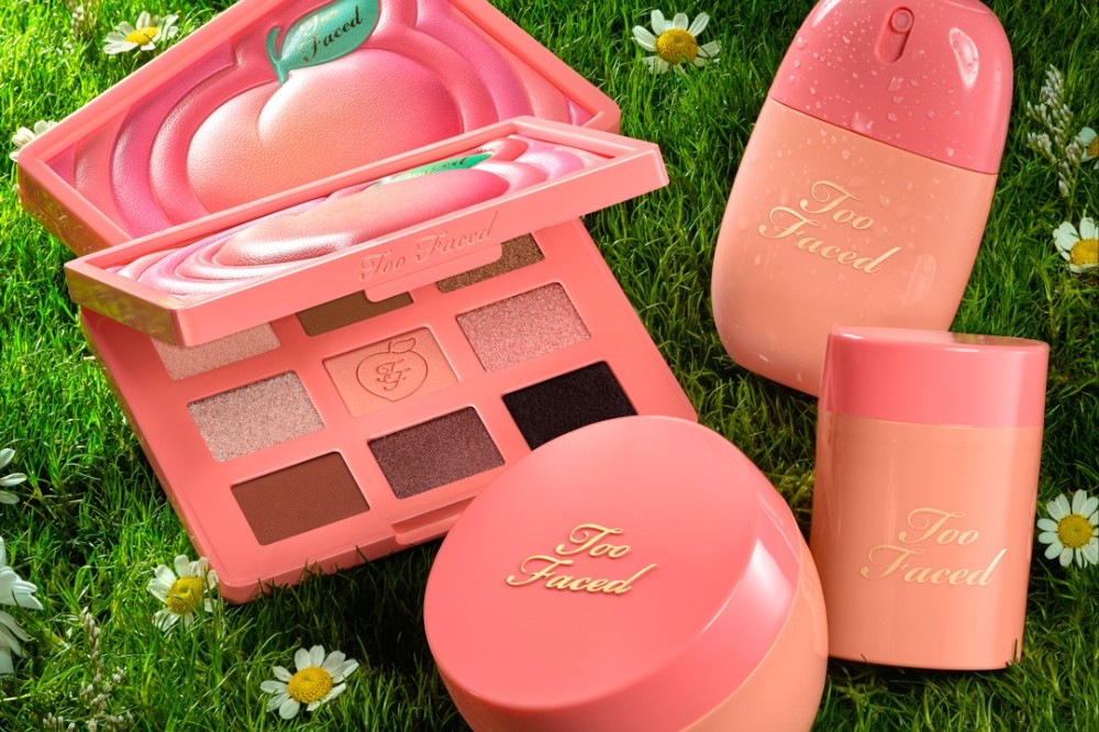 Produtos de maquiagem Too Faced em tons de pêssego e rosa, incluindo uma paleta de sombras aberta, um spray fixador e dois potes, dispostos sobre grama verde com margaridas brancas