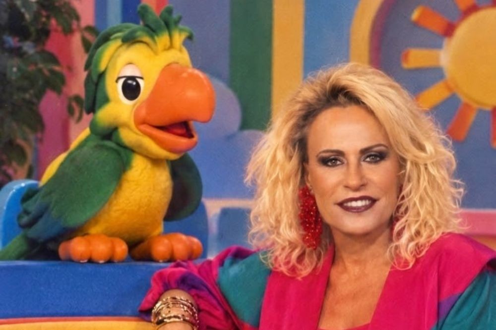 Apresentadora loira de cabelos cacheados e maquiagem forte, usando brincos vermelhos e blusa colorida, ao lado do boneco Louro José, um papagaio verde e amarelo com bico laranja, em um cenário de estúdio colorido