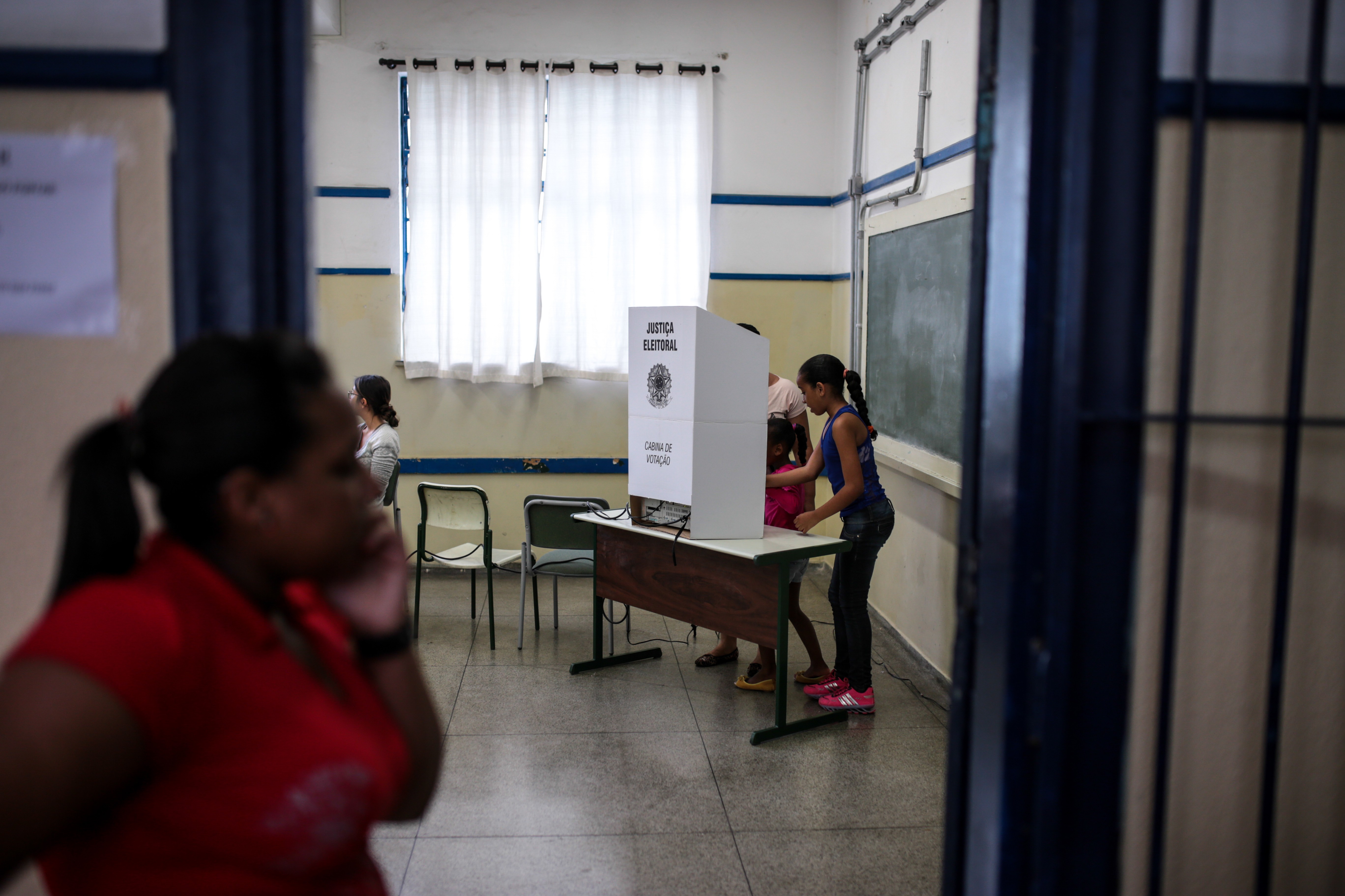 Duas crianças votando em uma urna eletrônica branca com o texto Justiça Eleitoral em uma sala de aula com paredes brancas e faixas azuis, cadeiras vazias e uma lousa. Uma mulher de blusa vermelha está em primeiro plano, desfocada