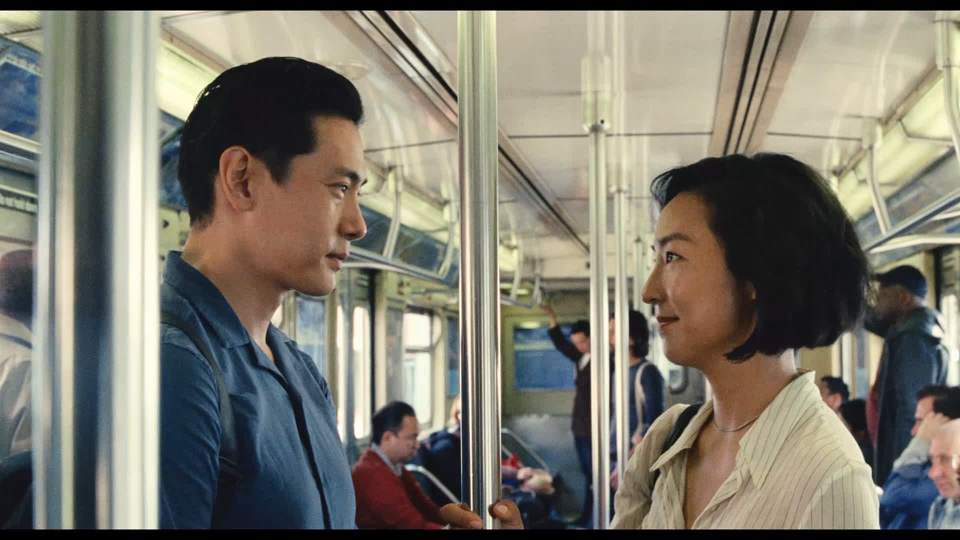 Um homem e uma mulher asiáticos se olham dentro de um trem. Ele, à esquerda, veste camisa azul e ela, à direita, camisa listrada clara, ambos sorrindo levemente. Barras verticais prateadas os separam, com outros passageiros desfocados ao fundo