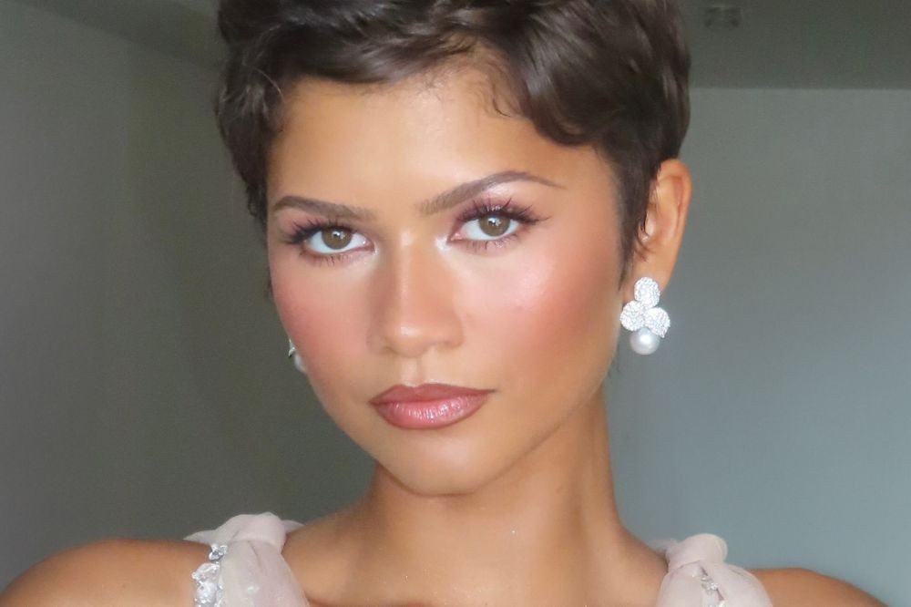 Close-up de Zendaya com cabelo curto e ondulado, maquiagem rosa suave, lábios brilhantes e brincos de pérola e diamante. Ela olha diretamente para a câmera com expressão séria