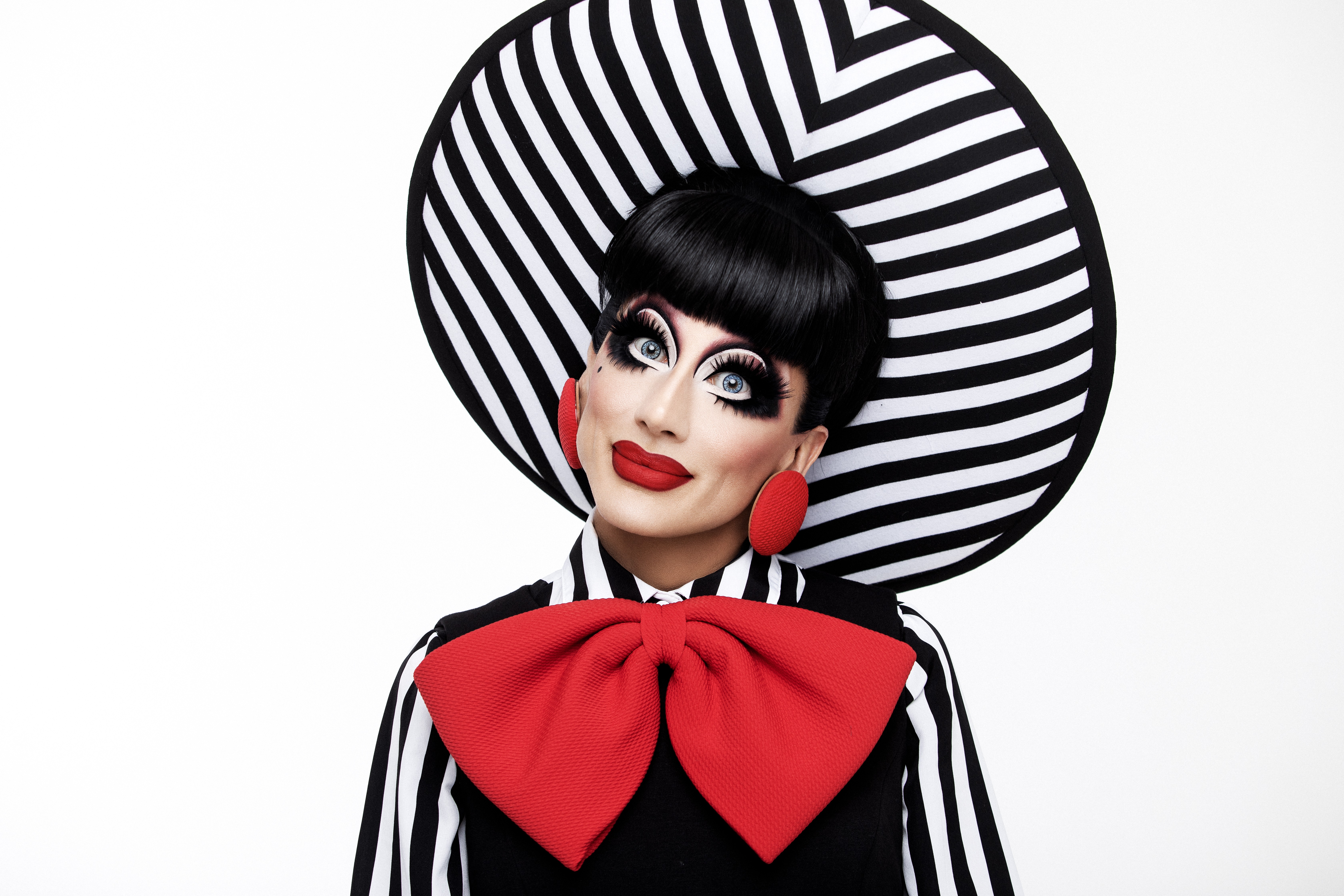 Drag queen com maquiagem dramática, batom vermelho e peruca preta. Usa um chapéu grande listrado em preto e branco, brincos vermelhos ovais e uma gravata borboleta vermelha gigante sobre uma camisa listrada