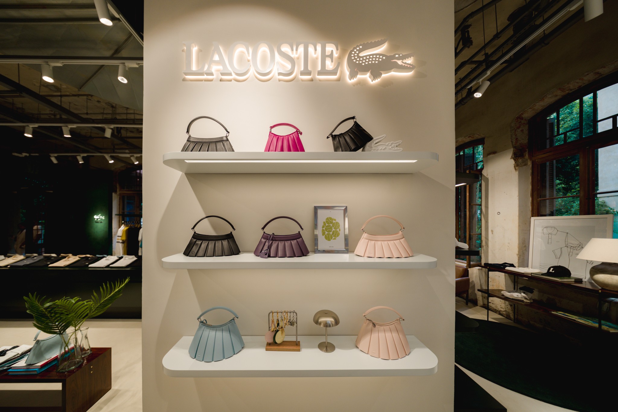 Prateleiras brancas com nove bolsas plissadas em tons de cinza, rosa, roxo, azul e nude, sob um letreiro neon da Lacoste com o logo do crocodilo. Ao fundo, um ambiente de loja com roupas e janelas grandes