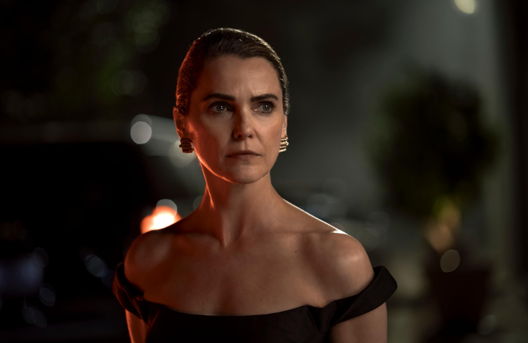 Mulher de pele clara, cabelos loiros presos, vestindo um elegante vestido preto tomara que caia e brincos dourados, olhando para o lado com expressão séria, em ambiente noturno com luzes desfocadas ao fundo