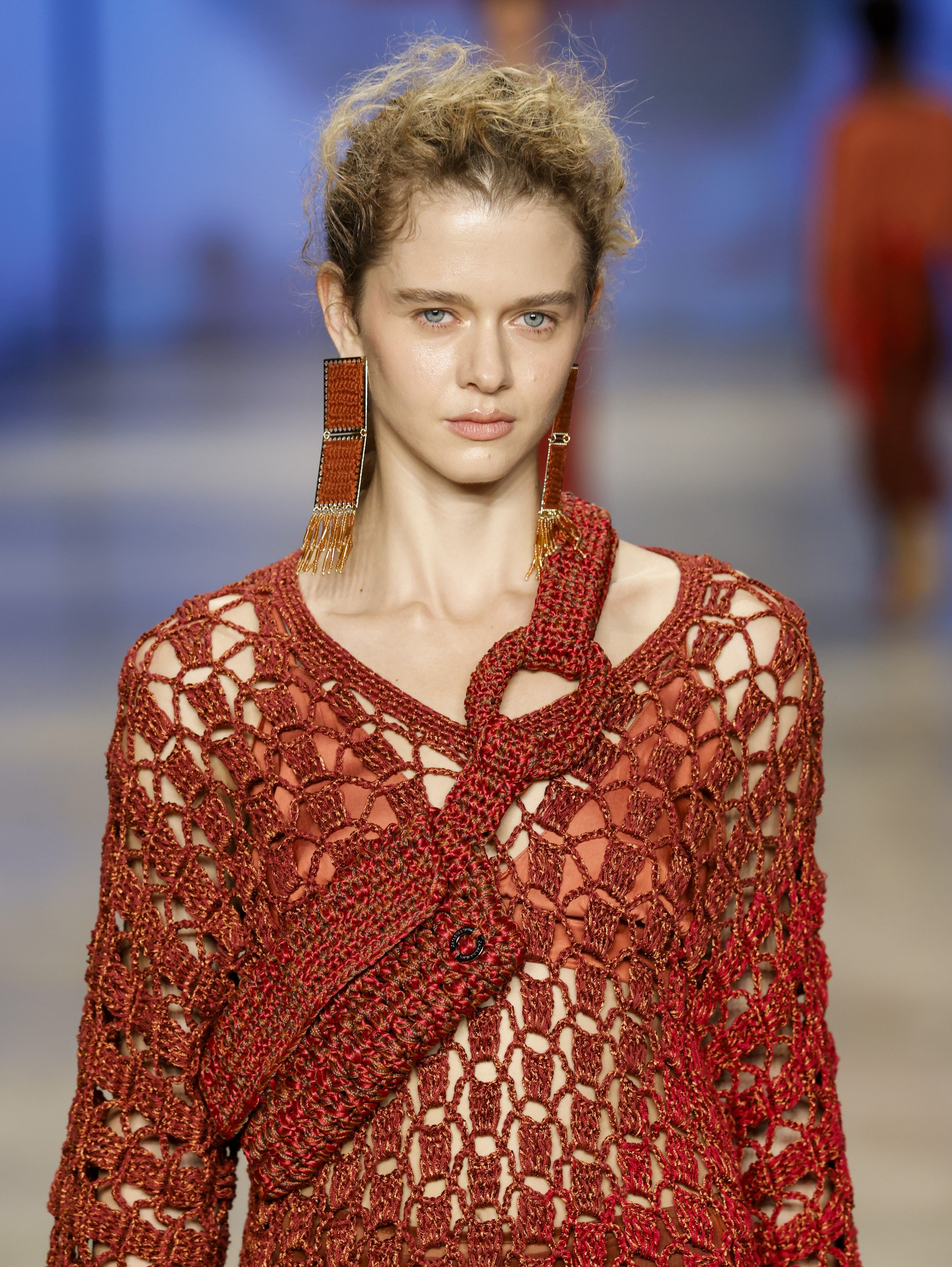 Modelo com cabelo loiro encaracolado preso, usando blusa de crochê marrom-avermelhada sobre top laranja e brincos longos retangulares com franjas douradas.
