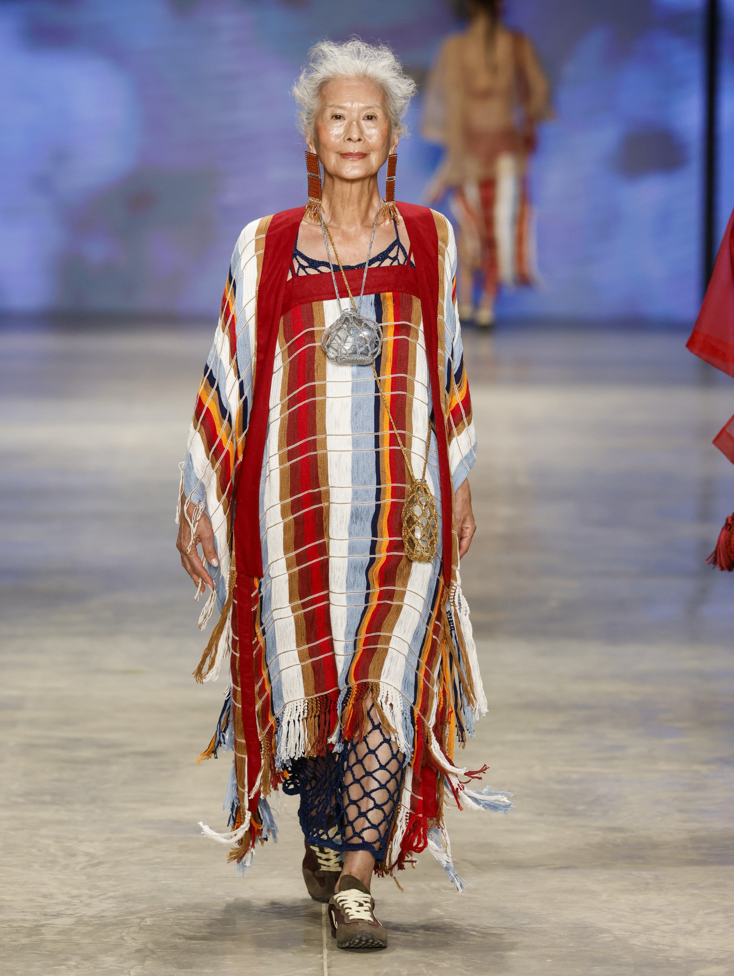 Mulher idosa com cabelos brancos e sorriso discreto desfila na passarela, vestindo um longo vestido xadrez colorido com franjas e detalhes em crochê azul, brincos longos e colares com pingentes grandes. Ela usa tênis marrons e caminha em direção ao observador