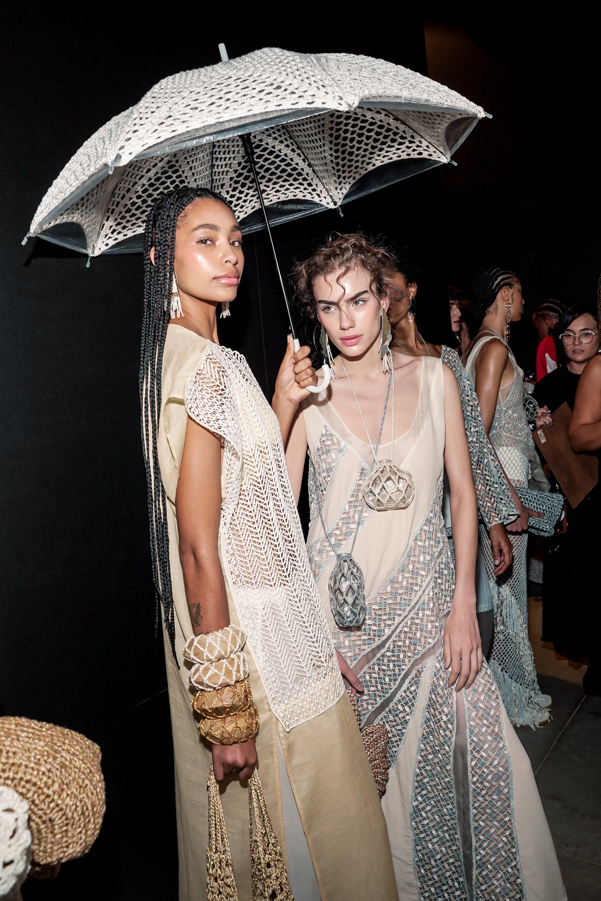 Duas modelos posam sob um guarda-chuva de crochê branco. A da esquerda, com tranças longas, veste túnica bege e pulseiras artesanais. A da direita, com cabelo cacheado, usa vestido bege com detalhes geométricos azuis e colares com pingentes de crochê. Outras modelos e pessoas desfocadas estão ao fundo escuro