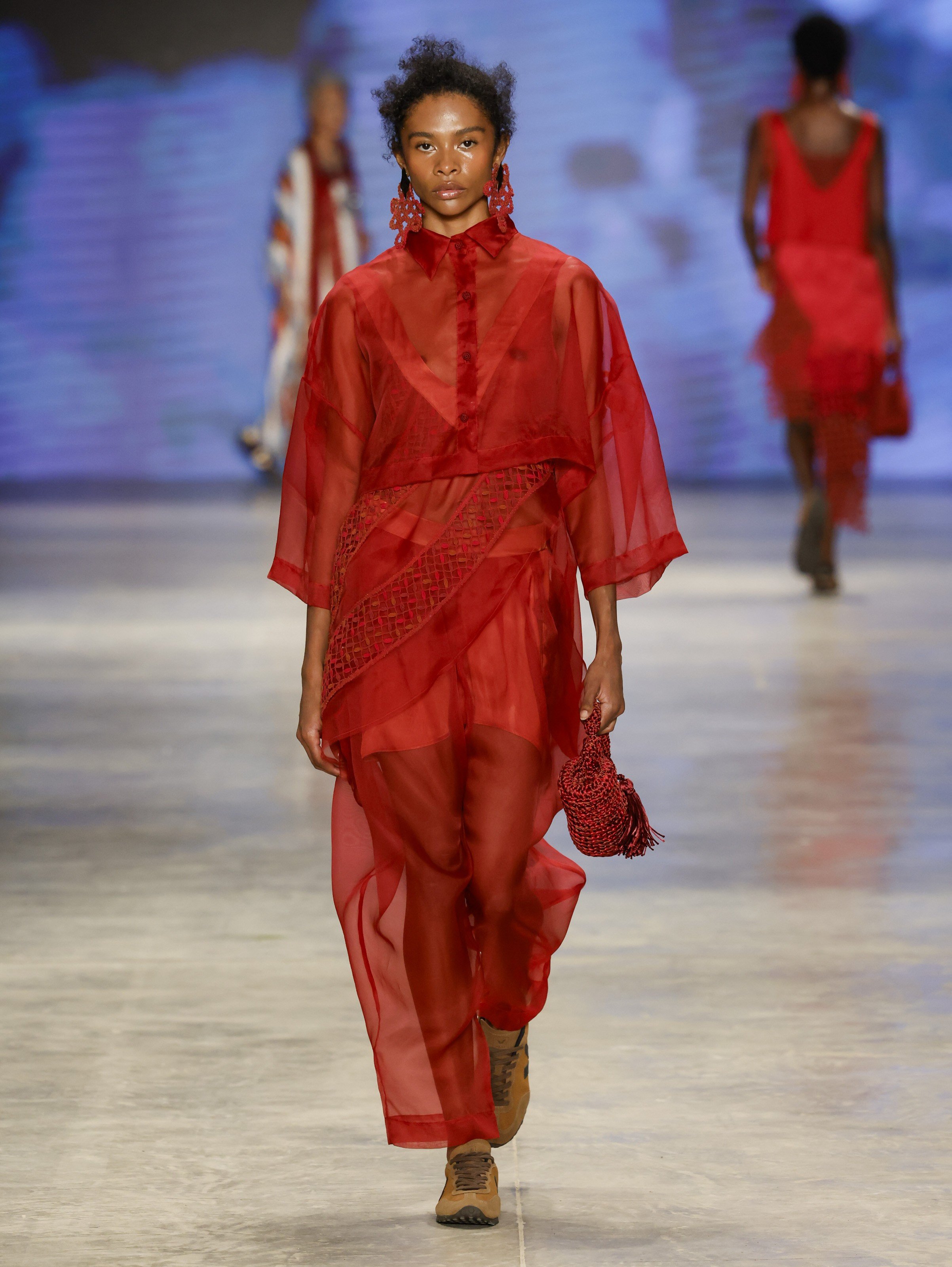 Modelo de pele escura e cabelo crespo curto, vestindo roupa vermelha transparente com calça e camisa de manga larga, brincos grandes vermelhos, bolsa de mão vermelha e sapatos marrons, desfilando em passarela cinza com fundo azul