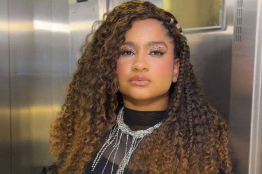 Mulher jovem de pele morena, cabelo castanho escuro e encaracolado, com tranças finas, usando maquiagem brilhante nos olhos e um colar prateado com franjas, posando em frente a uma parede metálica