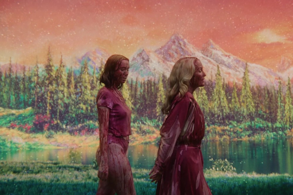 Duas mulheres, uma morena e outra loira, ambas com roupas e corpos manchados de vermelho, caminham em um cenário surreal de montanhas nevadas, florestas verdes e um lago, sob um céu alaranjado com estrelas