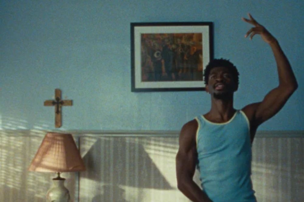Homem negro de regata azul clara com bordas amarelas, sorrindo e levantando o braço direito, em um quarto com parede azul, cruz de madeira, abajur e quadro na parede