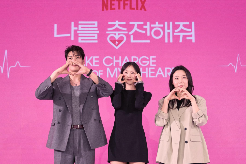 Três pessoas asiáticas sorrindo, em pé, fazendo gestos com as mãos em frente a um painel rosa com o logo da Netflix e texto em coreano e inglês. Um homem à esquerda, de terno cinza, faz um coração com as mãos. No centro, uma mulher de vestido preto faz corações com os dedos nas bochechas. À direita, outra mulher de blazer bege faz um coração com as mãos. O painel tem um eletrocardiograma branco