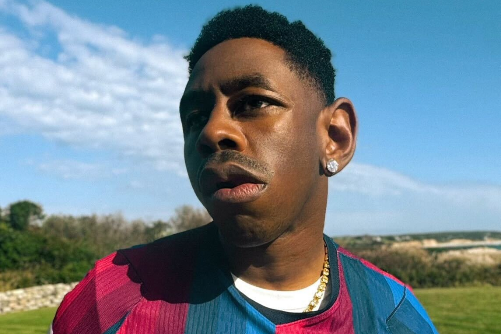 Homem negro com cabelo curto e crespo, usando brinco de diamante na orelha direita e colar dourado, veste camisa listrada azul e vermelha. Ele tem uma expressão séria, com a boca ligeiramente aberta, e olha para a esquerda. O fundo é um céu azul com nuvens esparsas e vegetação verde