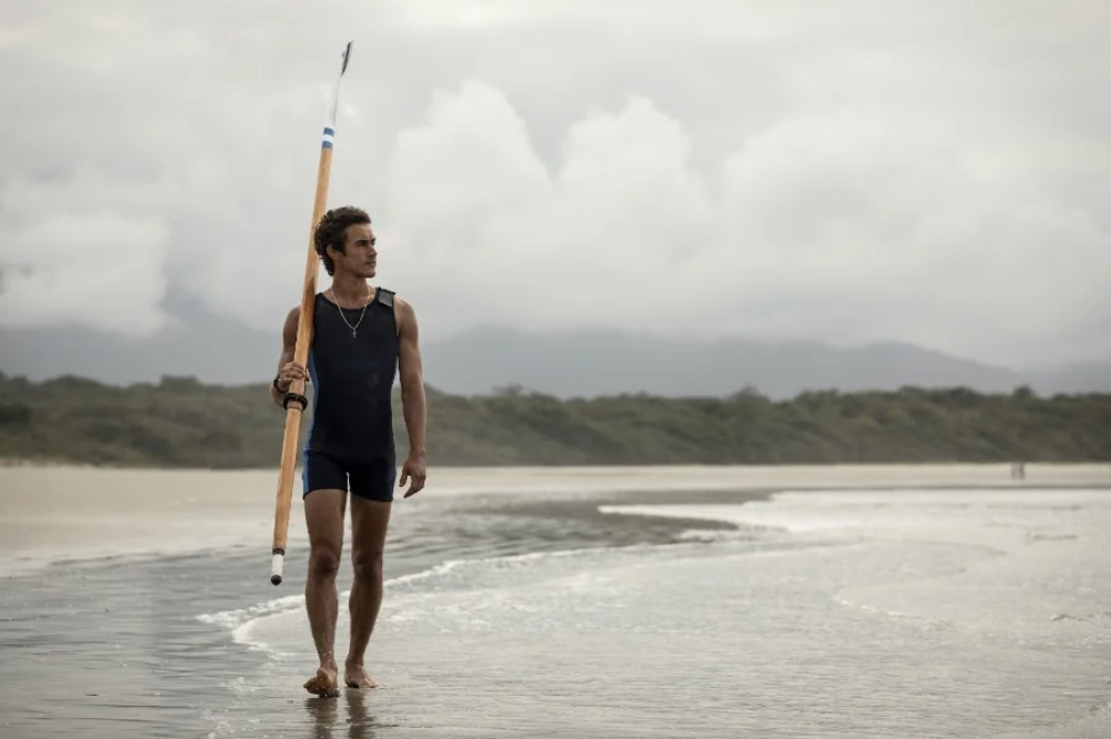Homem jovem, de pele clara e cabelos escuros, vestindo um uniforme de remo azul-marinho, caminha descalço pela beira da praia. Ele segura um remo de madeira com a mão direita, apoiado no ombro. O céu está nublado, e ao fundo há uma faixa de vegetação e montanhas.