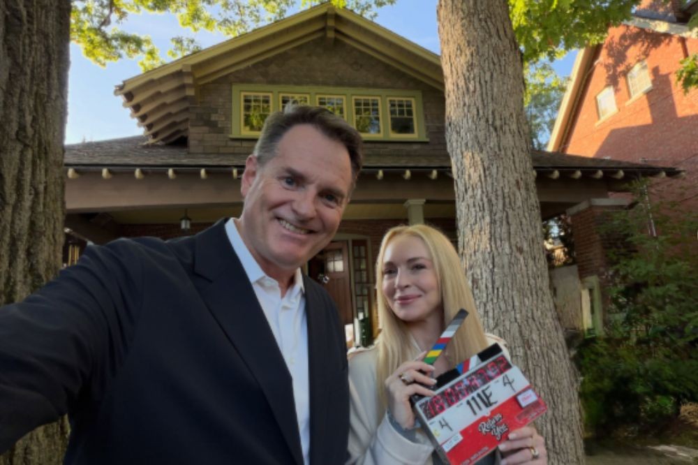 Homem sorridente de terno e mulher loira segurando uma claquete, ambos posando para selfie em frente a uma casa com árvores ao redor