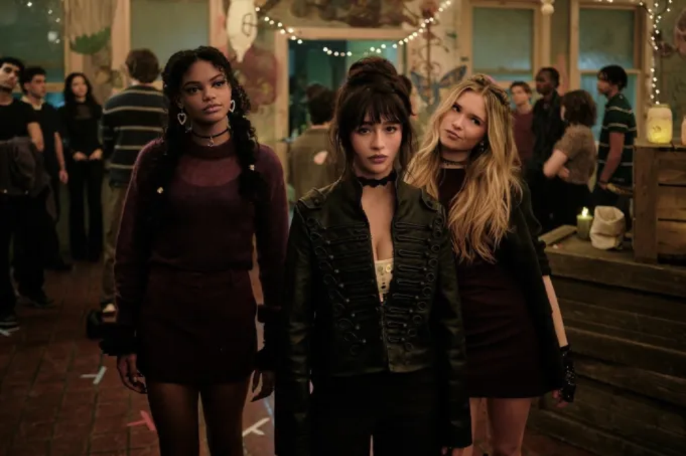 Três jovens mulheres com roupas escuras e colares choker, em pé, olhando para frente em uma festa com luzes e pessoas ao fundo. A da esquerda tem tranças e brincos de coração, a do centro usa jaqueta militar preta e a da direita tem cabelos loiros soltos e expressão de desdém
