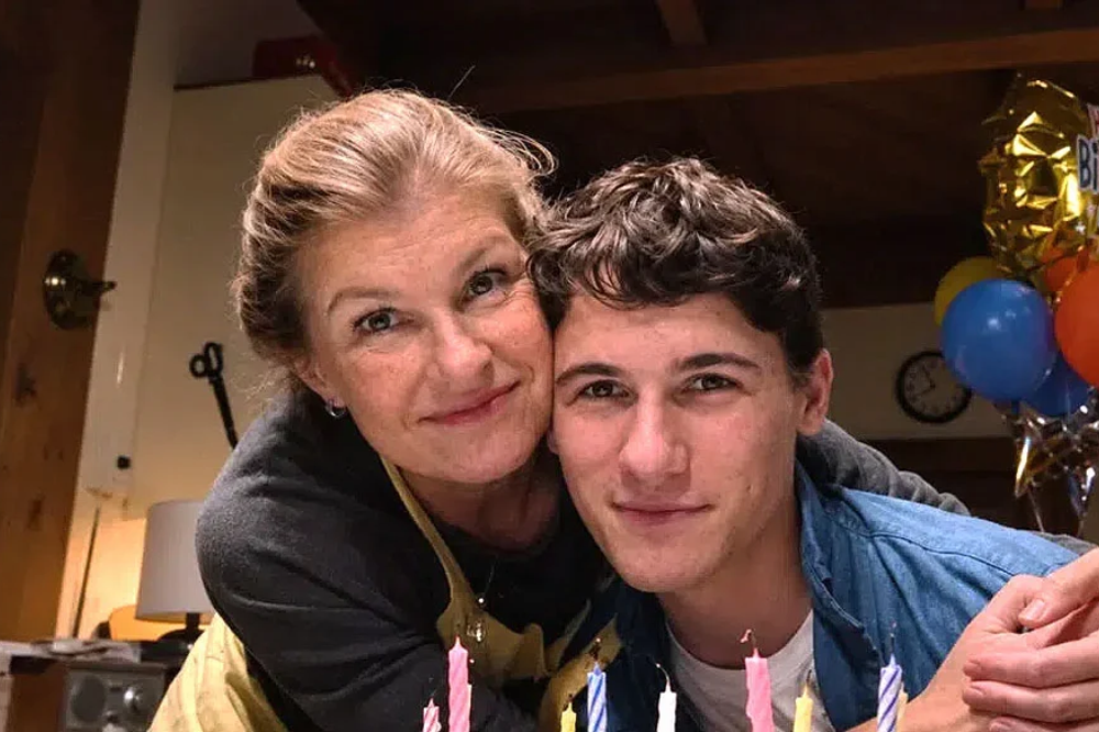 Mulher de cabelos loiros abraça um jovem de cabelos escuros, ambos sorrindo para a câmera. Velas de aniversário coloridas e balões festivos ao fundo.