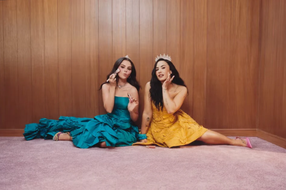 Selena Gomez e Demi Lovato sentadas no chão, usando coroas e vestidos de gala. Selena, à esquerda, veste azul-petróleo e segura um pincel de maquiagem. Demi, à direita, veste amarelo e toca o rosto. Ambas têm cabelos escuros e longos, e o fundo é uma parede de madeira com piso rosa.