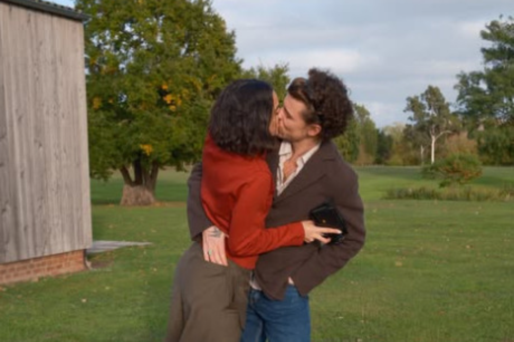 Casal se beijando apaixonadamente em um campo verde, com uma construção de madeira à esquerda e árvores ao fundo. A mulher, de cabelo escuro, veste blusa vermelha e saia verde-oliva. O homem, de cabelo encaracolado, usa jaqueta marrom, camisa clara e calça jeans, segurando uma carteira preta.