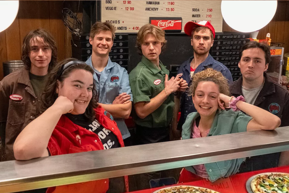 Sete jovens, cinco homens e duas mulheres, posam sorrindo atrás de um balcão de pizzaria. Duas pizzas estão em primeiro plano. Ao fundo, um cardápio com preços e um logo da Coca-Cola.