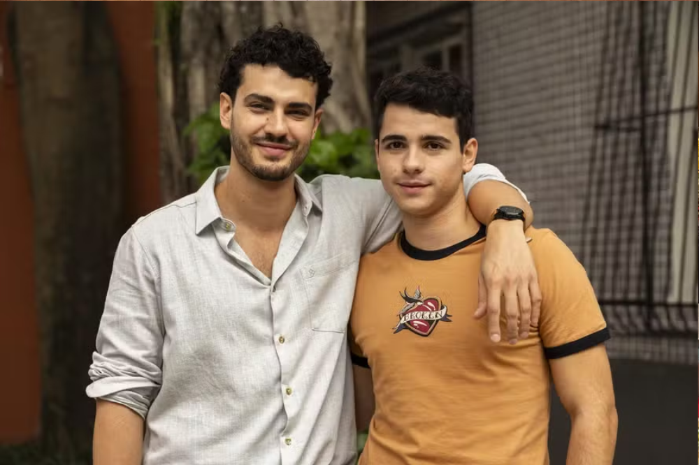 Dois homens jovens sorrindo para a câmera, o da esquerda com barba e camisa cinza clara, o da direita com camiseta mostarda e braço do outro homem sobre seu ombro, em ambiente externo com folhagem e parede marrom ao fundo