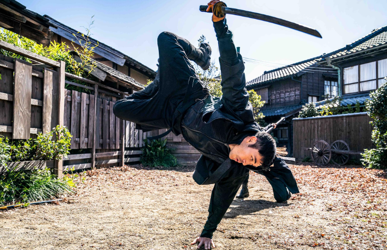 Homem asiático de roupa preta, com uma katana na mão direita, realizando um movimento acrobático com o corpo inclinado e uma mão no chão, em um pátio de terra batida com casas japonesas e cercas de madeira ao fundo