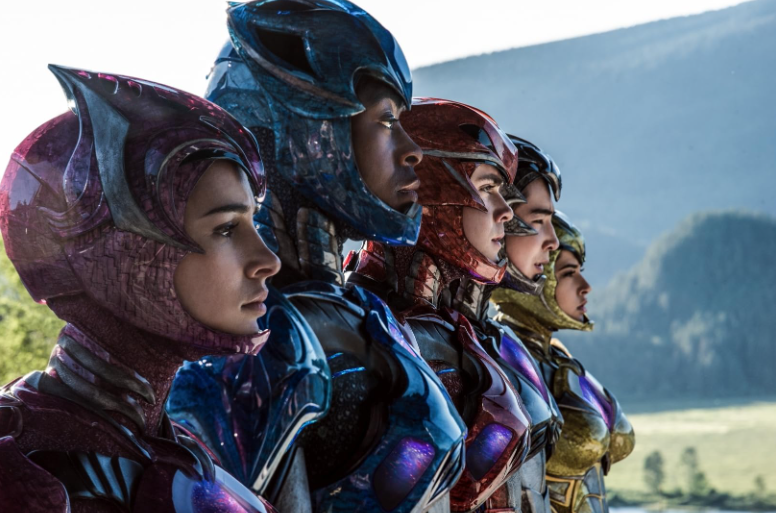 Cinco jovens, os Power Rangers, perfilados da esquerda para a direita, com capacetes e armaduras coloridas: rosa, azul, vermelho, amarelo e preto. Eles olham para o horizonte em um cenário montanhoso e natural