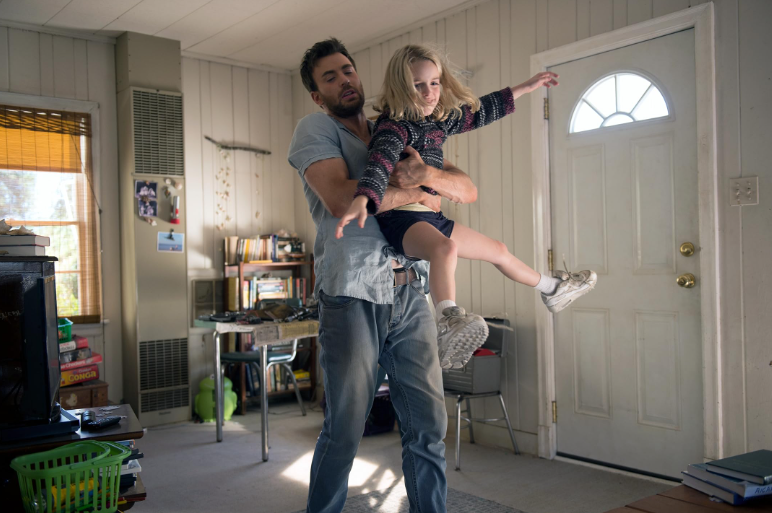 Um homem barbudo de camisa azul clara e calça jeans segura uma menina loira de suéter colorido e shorts escuros, que estende os braços e uma perna como se estivesse voando. Eles estão em uma sala com paredes de madeira clara, uma janela com persiana, uma porta branca e estantes de livros ao fundo