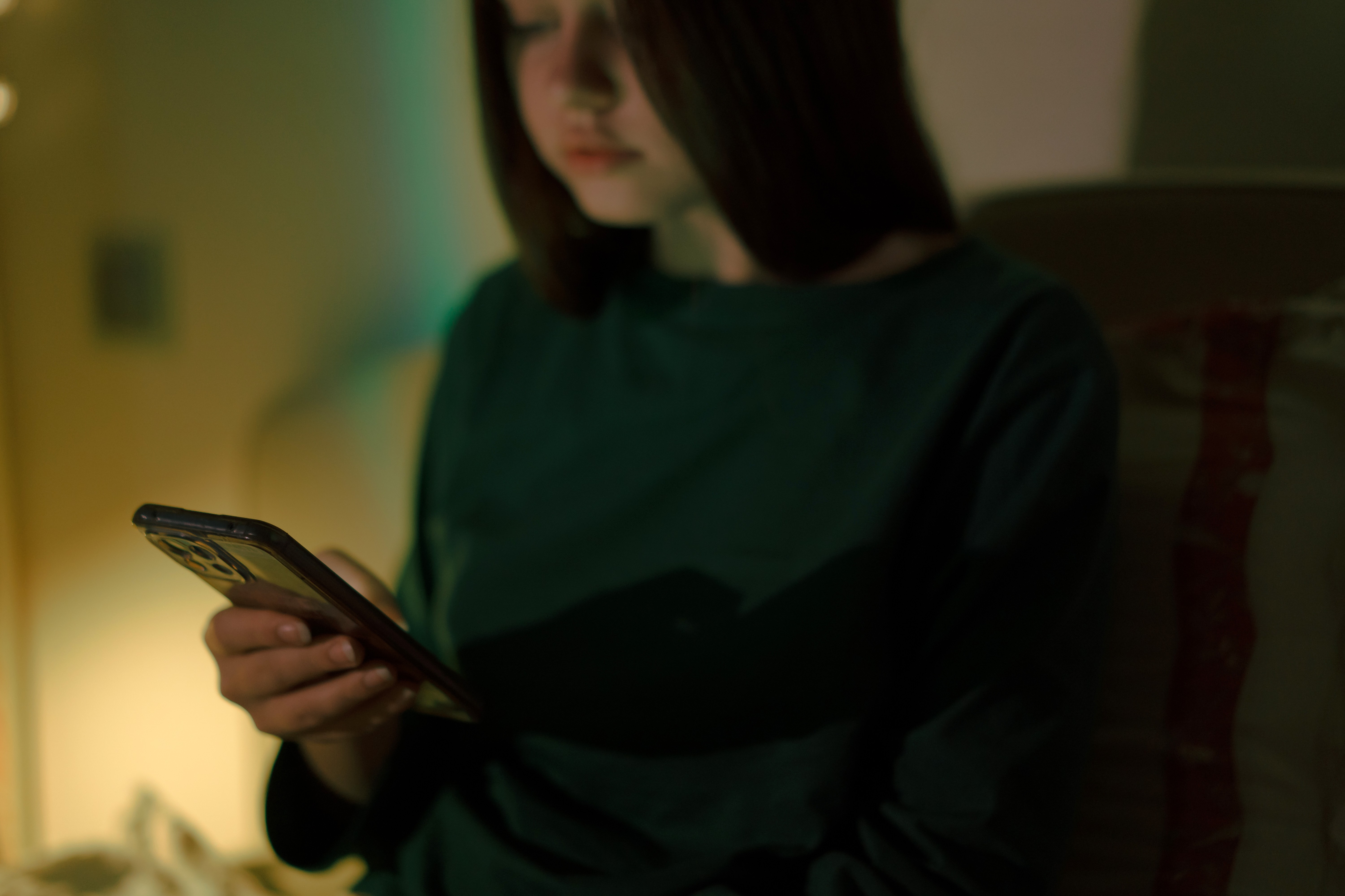 Jovem de cabelos longos e escuros, vestindo blusa verde-escura, olhando para um smartphone com a tela iluminada, em ambiente com pouca luz e tons esverdeados