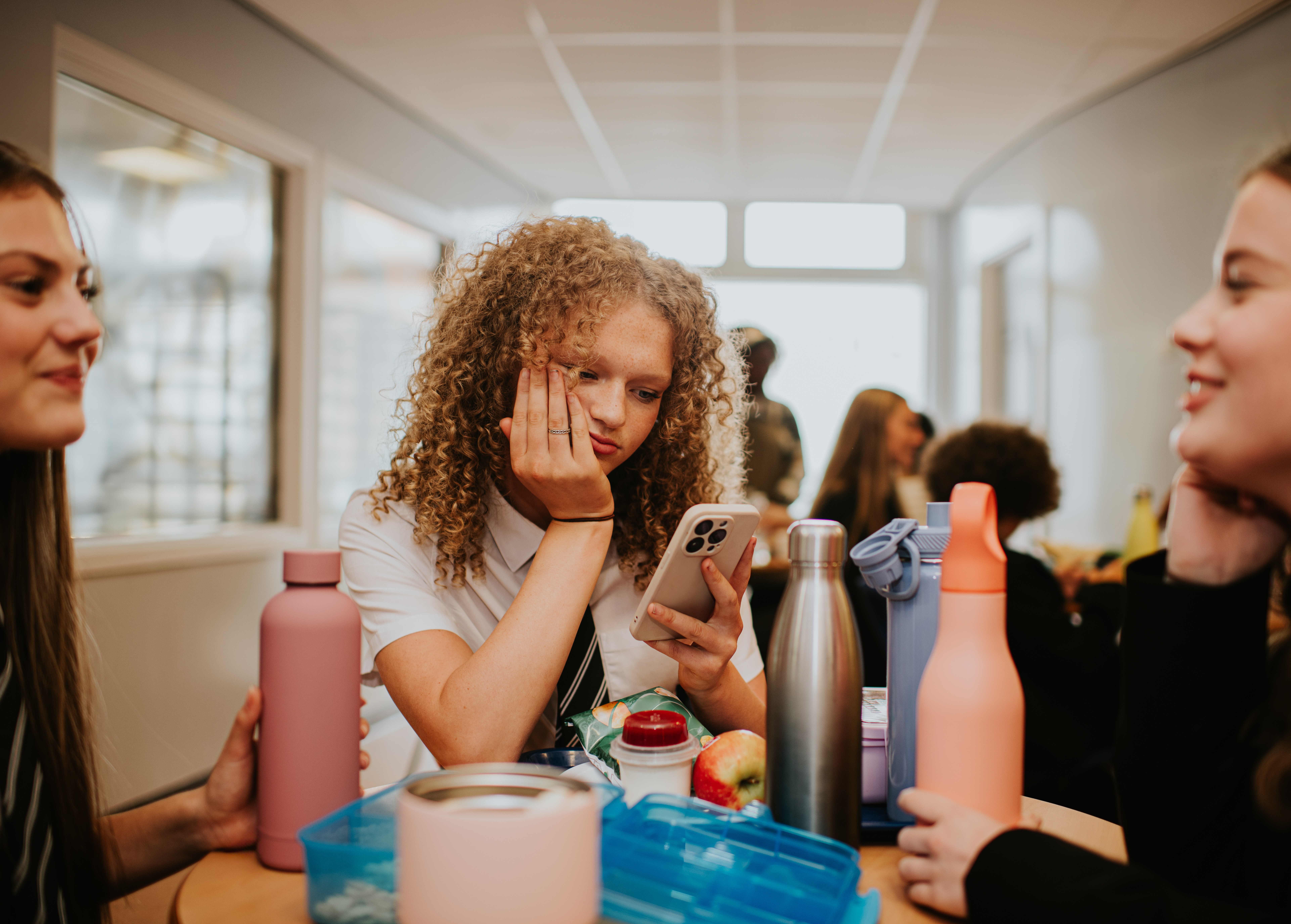 Três adolescentes em uma mesa de refeitório. A do centro, com cabelo cacheado, olha entediada para o celular, apoiando o rosto na mão. Garrafas de água coloridas e lanches estão sobre a mesa. As outras duas sorriem, conversando entre si