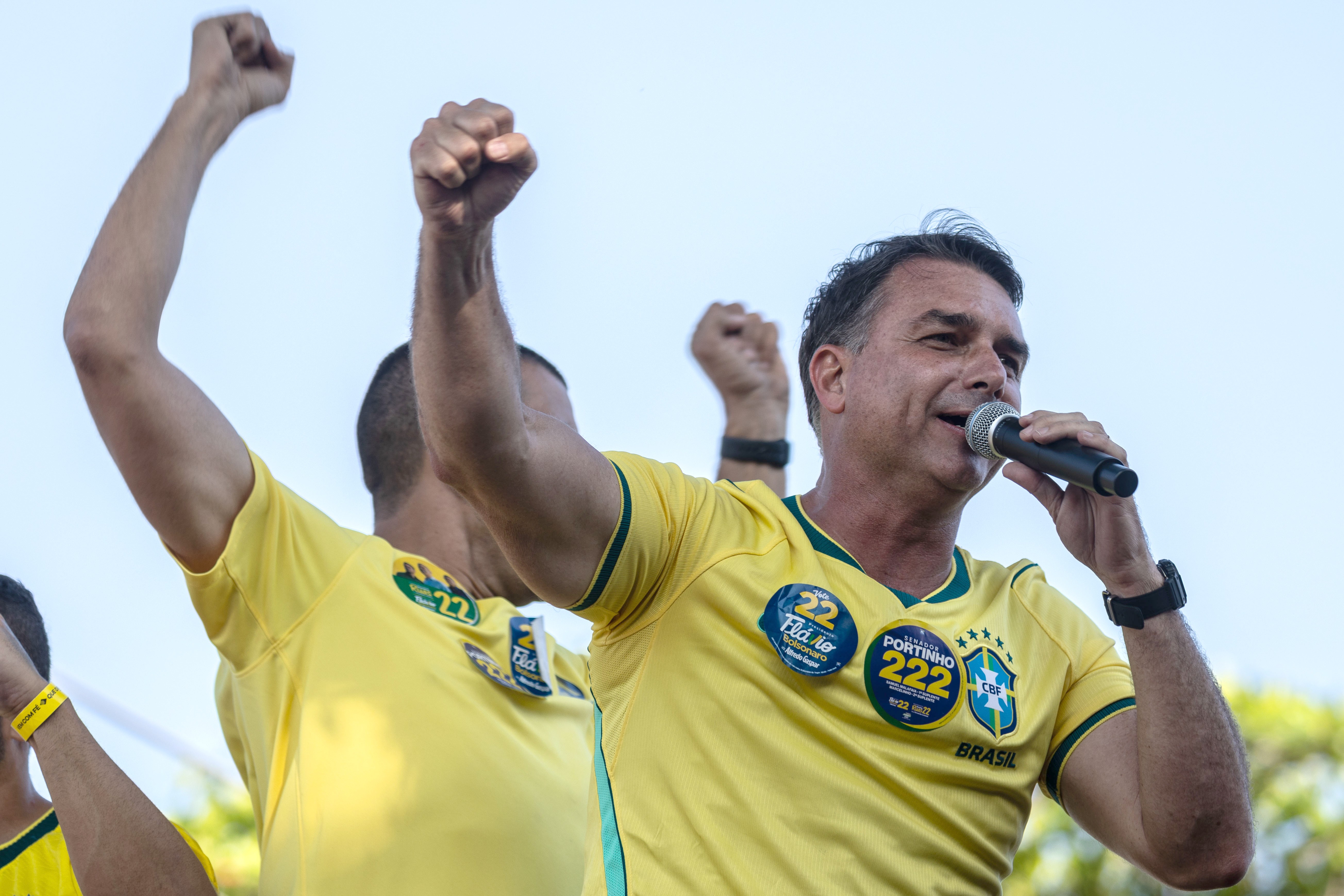 Jair Bolsonaro, sorrindo, veste camisa amarela da seleção brasileira com broches de campanha e segura um microfone. Ao fundo, outras pessoas com camisas amarelas levantam os punhos em sinal de comemoração