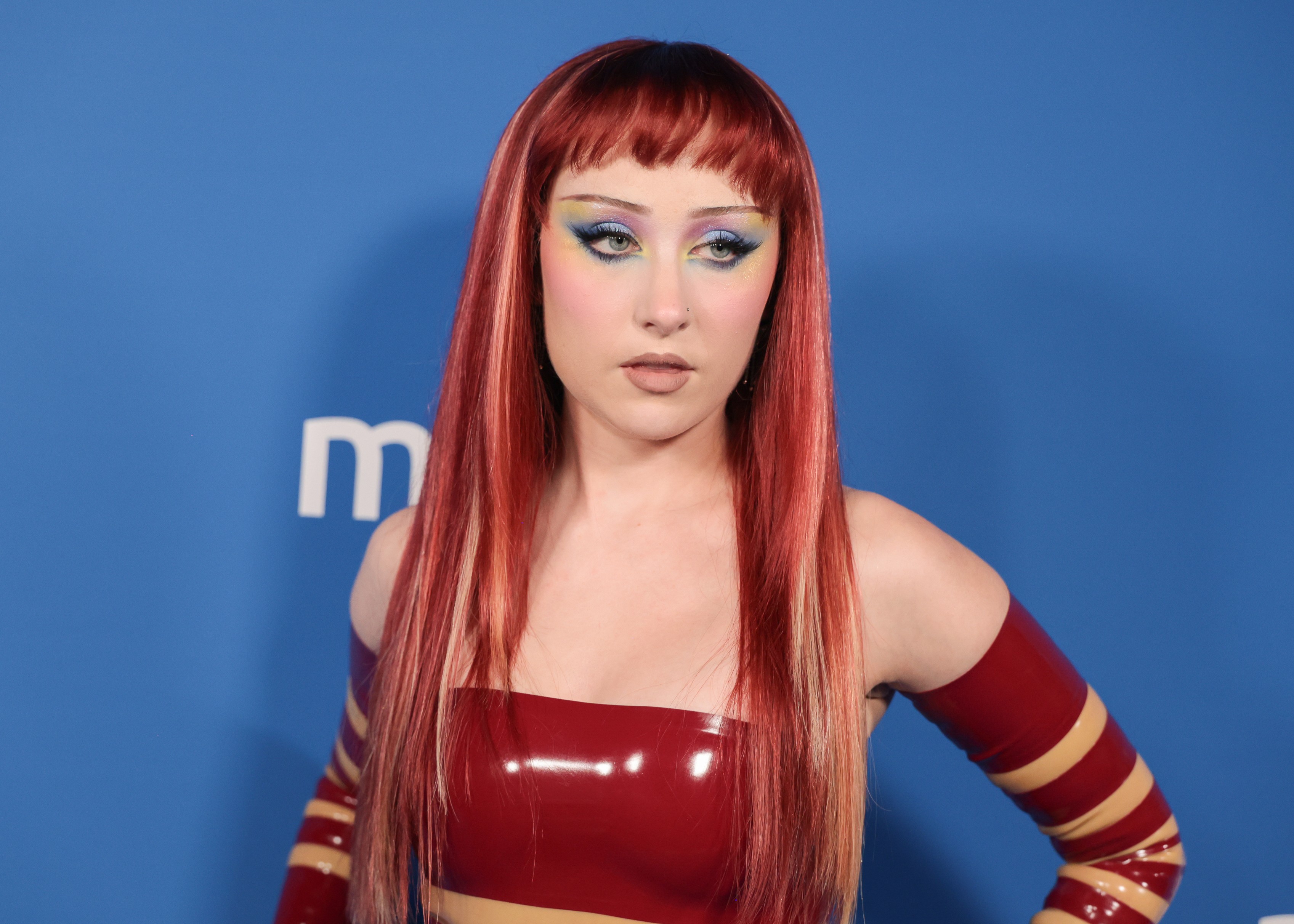 Cantora Pabllo Vittar com cabelo ruivo longo e franja curta, maquiagem colorida nos olhos e batom nude. Veste top tomara que caia e luvas vermelhas com listras amarelas, posando contra fundo azul