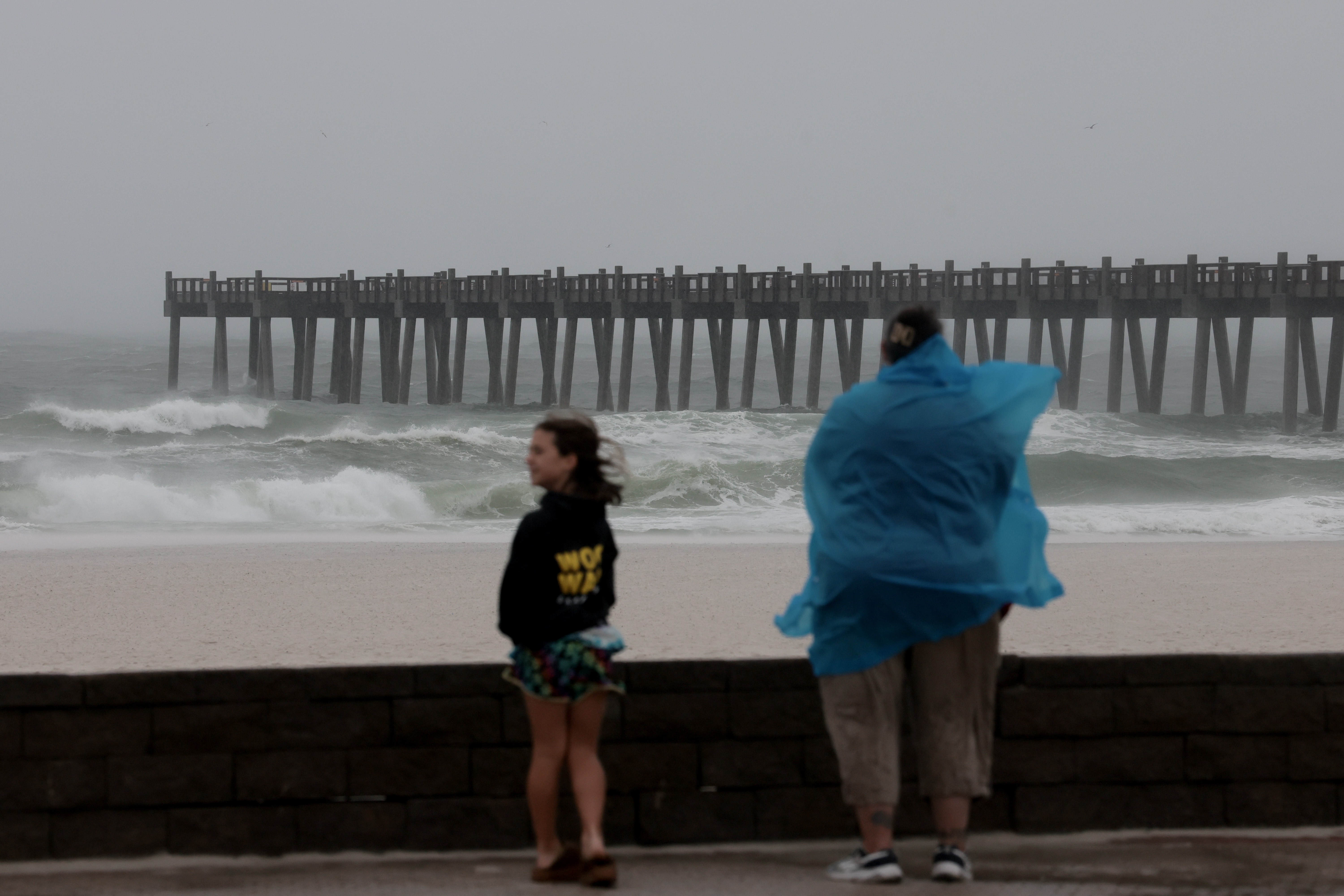 Duas pessoas observam o mar agitado em dia nublado, com um píer ao fundo. Uma menina de moletom preto e shorts floridos está à esquerda, e uma pessoa de costas com capa de chuva azul esvoaçante está à direita.