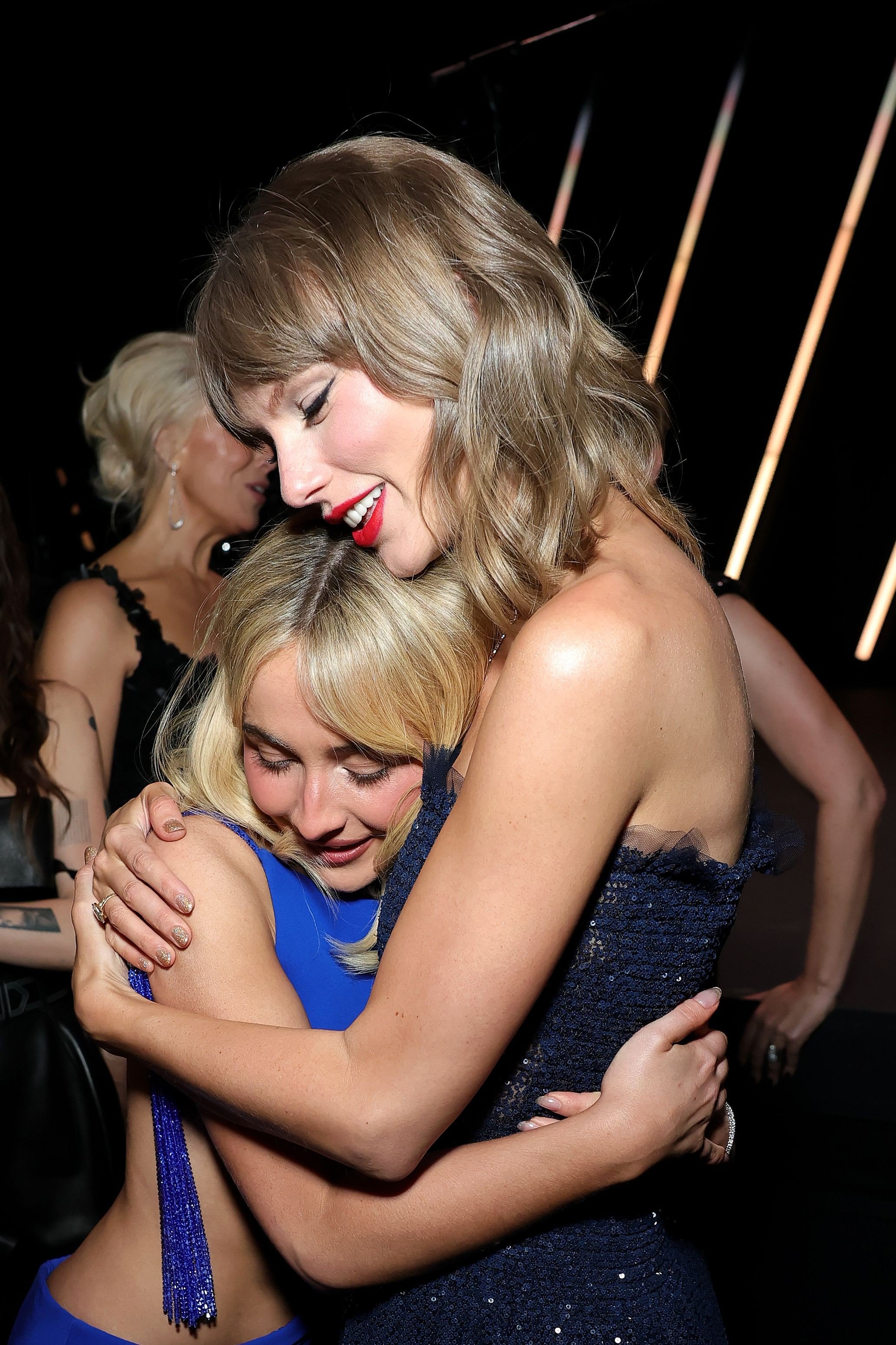 Taylor Swift, com cabelo loiro ondulado e batom vermelho, abraça Sabrina Carpenter, de cabelo loiro liso e vestido azul, em um evento noturno. Ambas sorriem com os olhos fechados, demonstrando carinho.