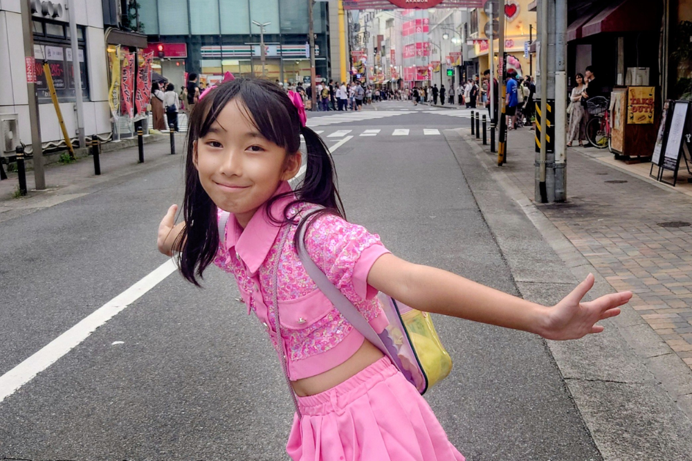 Menina asiática sorridente, com cabelo preto preso em duas marias-chiquinhas com laços rosa, vestindo um conjunto rosa brilhante de blusa cropped e saia, e uma mochila amarela transparente, posa com os braços abertos em uma rua movimentada com prédios e pessoas ao fundo