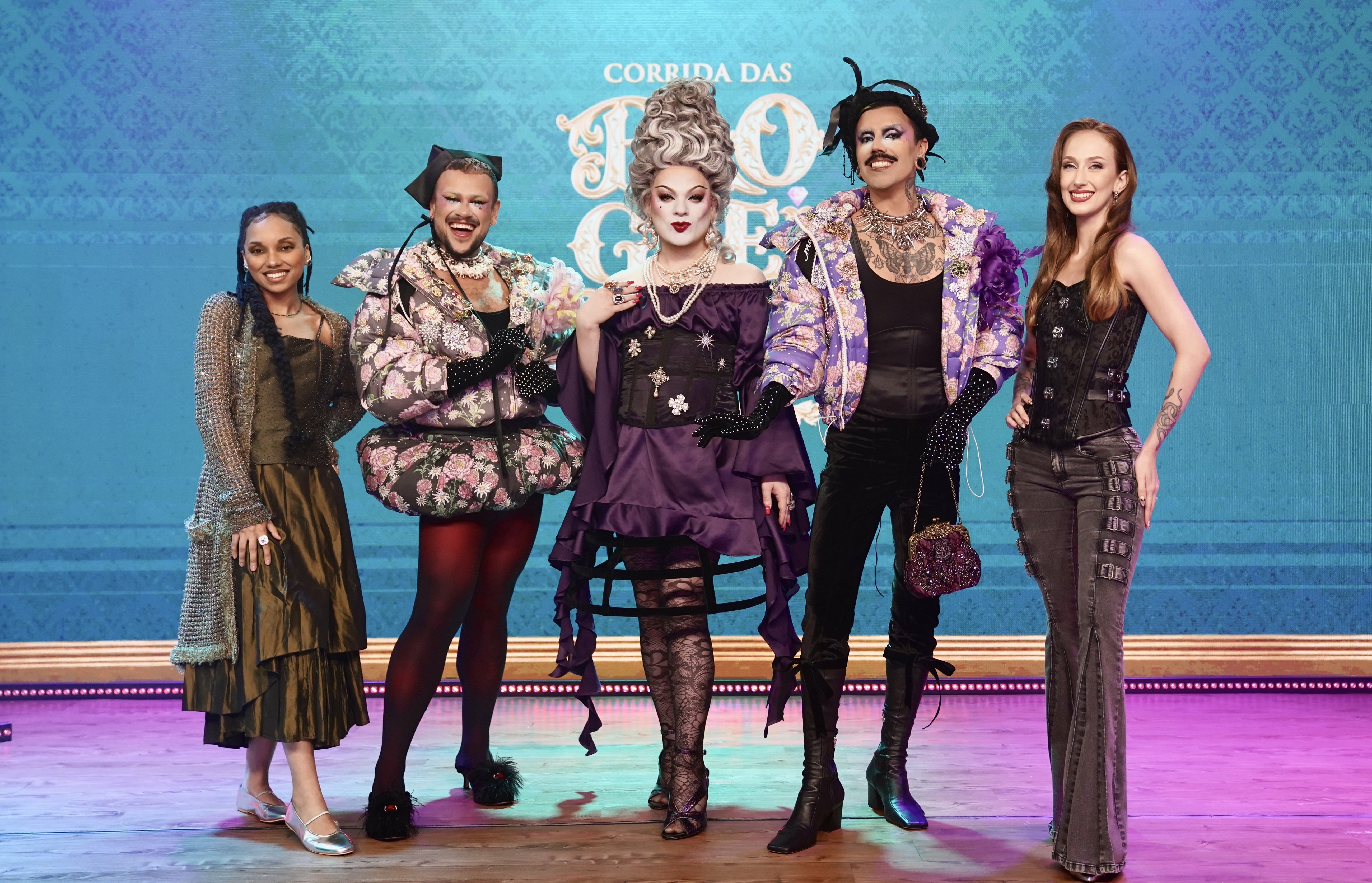 Cinco pessoas, incluindo três drag queens, posam sorridentes em um palco com fundo azul e o letreiro Corrida das Blogueiras. Da esquerda para a direita: uma mulher de vestido verde e casaco brilhante; uma drag queen de casaco floral rosa e preto, meia-calça vermelha; uma drag queen de peruca branca volumosa, vestido roxo e luvas pretas; uma drag queen de bigode, casaco roxo estampado e botas pretas; e uma mulher de espartilho preto e calça jeans escura