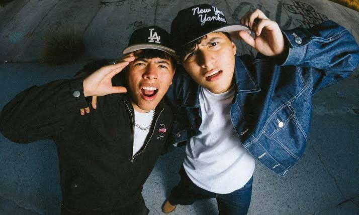 Dois jovens asiáticos, um à esquerda com boné LA e jaqueta preta, outro à direita com boné New York Yankees e jaqueta jeans, posam com expressões surpresas e bocas abertas, olhando para cima. O fundo é um muro cinza escuro com grafites