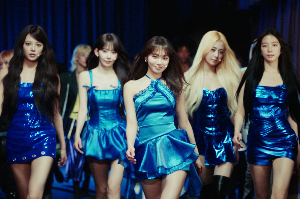 Cinco mulheres jovens, com cabelos longos e maquiagem leve, usam vestidos azuis brilhantes e caminham em fila, sorrindo levemente. A mulher do centro veste um vestido azul-turquesa com gola halter, enquanto as outras usam tons de azul mais escuros, alguns com lantejoulas. O fundo é um palco escuro com iluminação azul