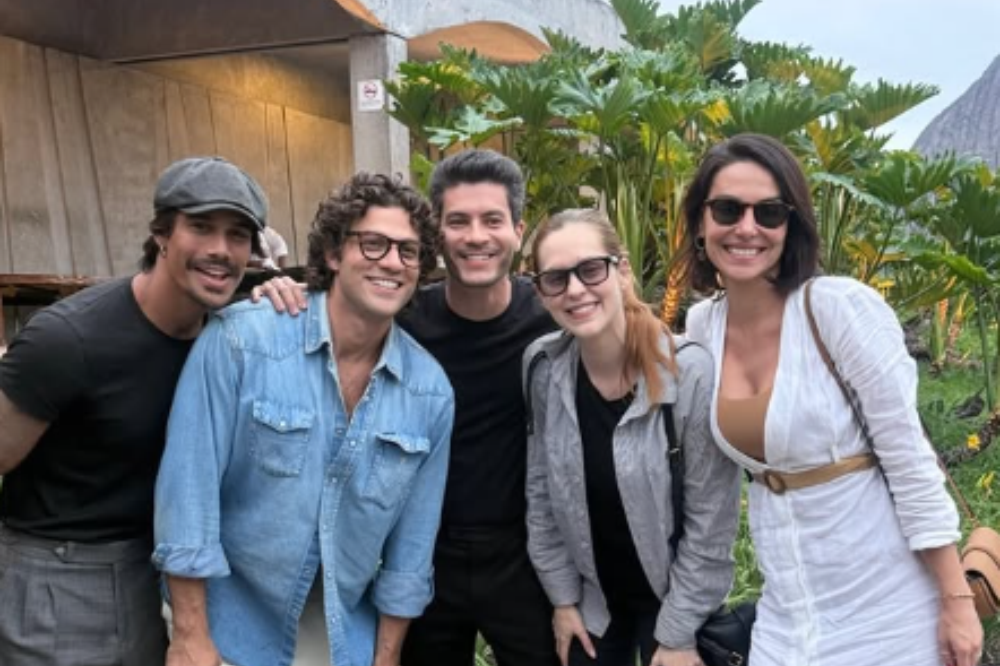 Cinco pessoas sorrindo para a câmera, com um homem de boné cinza e outro de camisa jeans à esquerda, um homem de camiseta preta no centro, uma mulher de óculos escuros e jaqueta cinza, e uma mulher de óculos escuros e vestido branco à direita. Ao fundo, folhagens verdes e uma montanha.