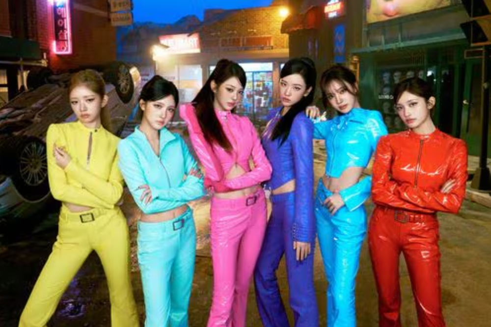 Seis mulheres jovens, com cabelos escuros e lisos, posam em uma rua noturna com um carro virado ao fundo. Elas vestem conjuntos de calça e jaqueta de couro brilhante, cada uma em uma cor diferente: amarelo, azul-claro, rosa, roxo, azul-turquesa e vermelho. Todas têm expressões sérias e braços cruzados ou mãos nos quadris