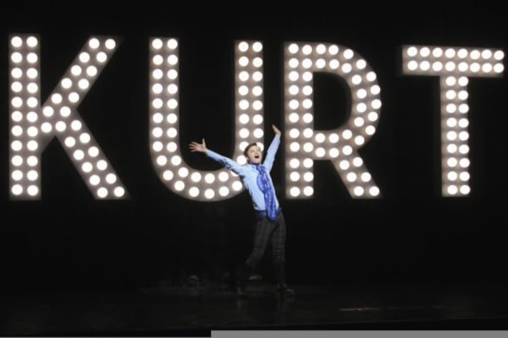 Jovem sorridente, de camisa azul e gravata estampada, braços abertos e corpo inclinado, em frente a um letreiro luminoso com o nome KURT em letras maiúsculas, sobre fundo escuro