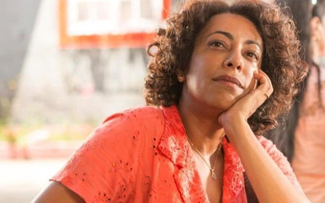 Mulher de pele morena e cabelos cacheados castanhos, vestindo blusa coral com detalhes vazados, apoia o queixo na mão direita, olhando para cima com expressão pensativa