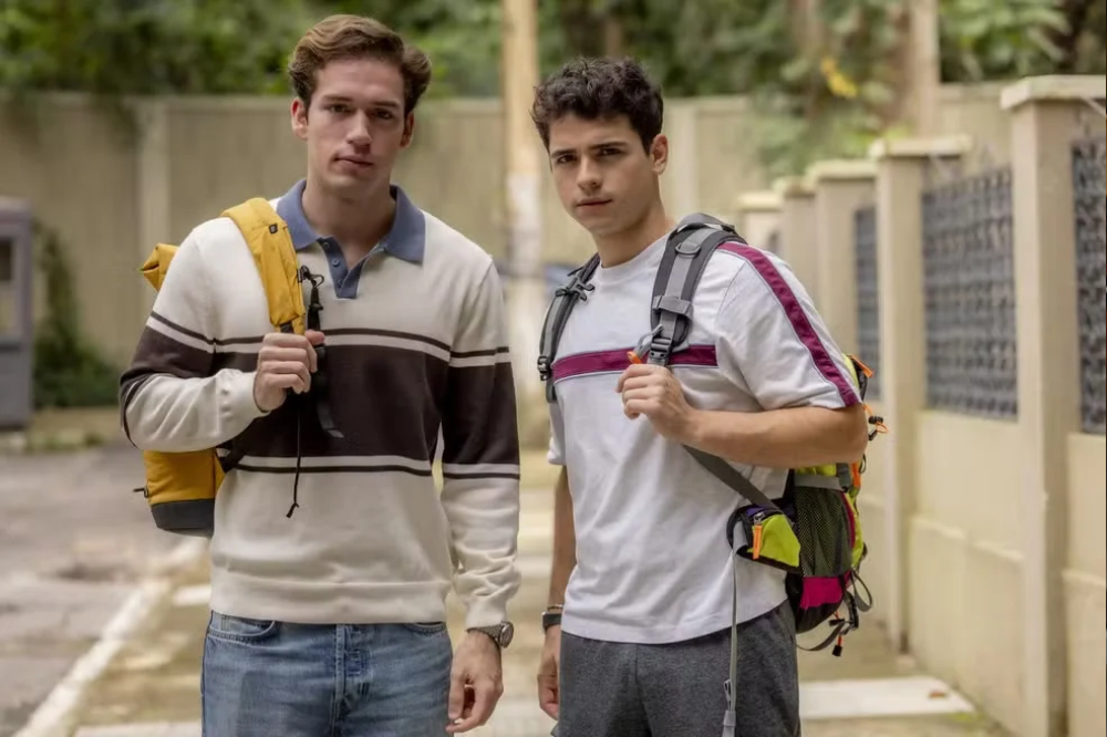 Dois jovens, um loiro e outro moreno, com mochilas, olham para frente. O loiro veste suéter listrado e jeans; o moreno, camiseta branca com faixa vinho e calça cinza. Estão em uma calçada com muro bege e vegetação ao fundo