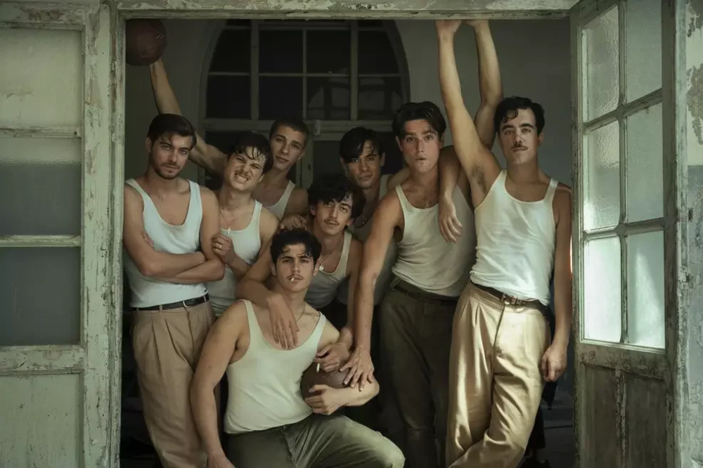 Grupo de nove jovens, a maioria com bigodes finos, vestindo regatas brancas e calças de cintura alta, posando em uma porta antiga. Um segura uma bola de futebol americano, outro um cigarro, e um terceiro levanta os braços com uma bola de basquete.