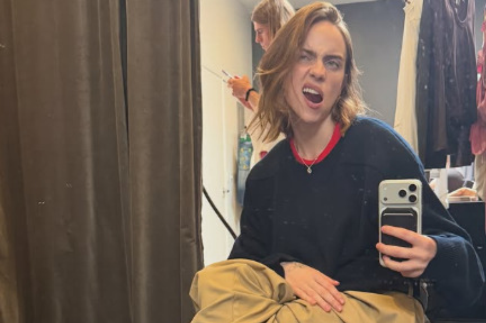 Jovem de cabelo castanho claro, com expressão de raiva e boca aberta, tira selfie em espelho. Veste suéter azul-marinho com gola vermelha, calça cáqui e colar prateado. Ao fundo, cortina marrom e arara de roupas.