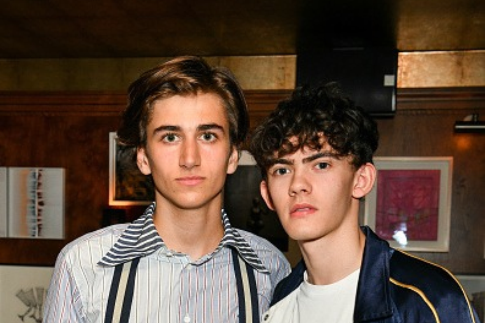 Dois jovens, um com cabelo liso e camisa listrada com suspensórios, e outro com cabelo cacheado e jaqueta azul, posam sérios em um ambiente interno com quadros na parede