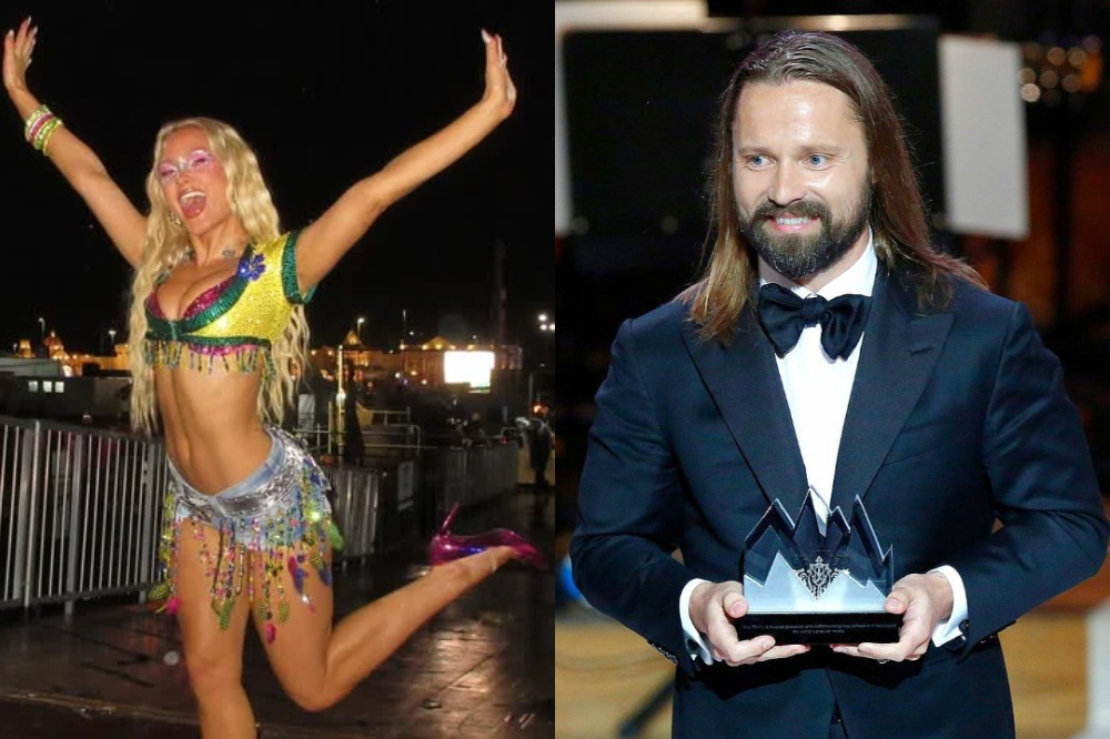 À esquerda, uma mulher loira sorridente, com top e shorts brilhantes, braços erguidos em celebração. À direita, um homem de cabelos longos e barba, vestindo terno e gravata borboleta, segurando um troféu de cristal