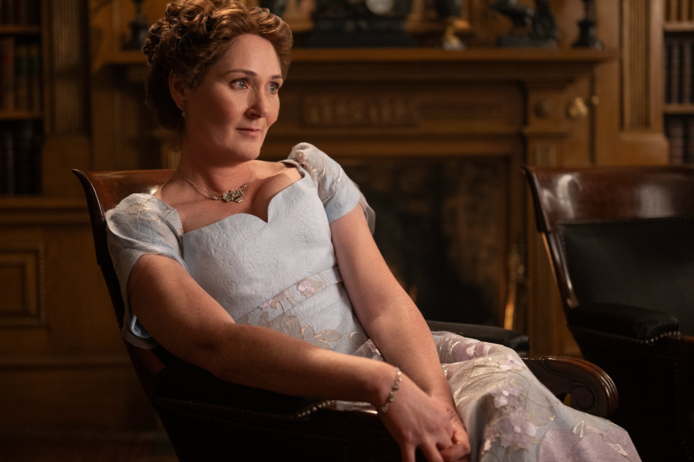 Mulher de meia-idade, cabelos castanhos presos, sentada em uma poltrona de couro, usando um vestido azul claro com detalhes florais e um colar dourado. Ela olha para a direita com uma expressão serena, em um ambiente com estante de livros e lareira ao fundo