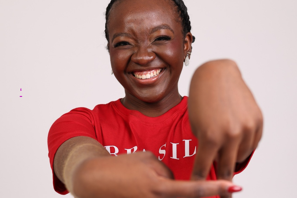 Mulher negra sorridente, com tranças e brincos prateados, vestindo camiseta vermelha com BRASIL em branco, apontando para baixo com as duas mãos, unhas vermelhas