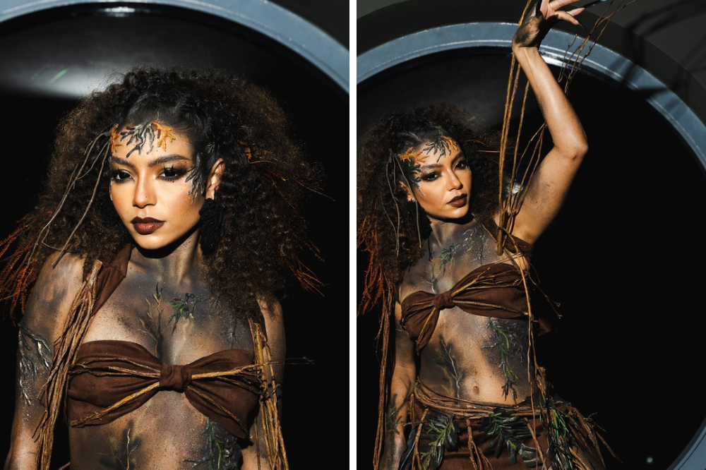 Mulher com maquiagem e figurino de natureza, cabelo cacheado volumoso, maquiagem escura nos olhos e galhos no rosto. Usa um top marrom amarrado na frente, com o corpo pintado de cinza e detalhes de folhas e galhos. Na imagem da direita, ela levanta um braço, segurando galhos finos, com um fundo escuro e um arco cinza ao redor